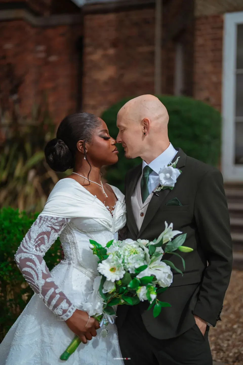 Helen and Jack kissing beside the brick house at Shottle Hall, with Helen holding her white bouquet