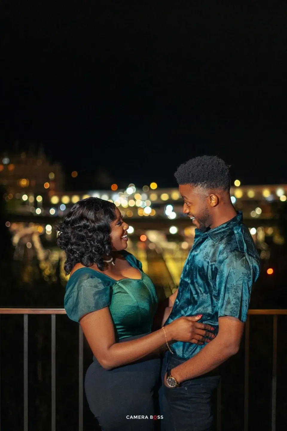 A tender nighttime couple portrait on a balcony with twinkling city lights in the background.