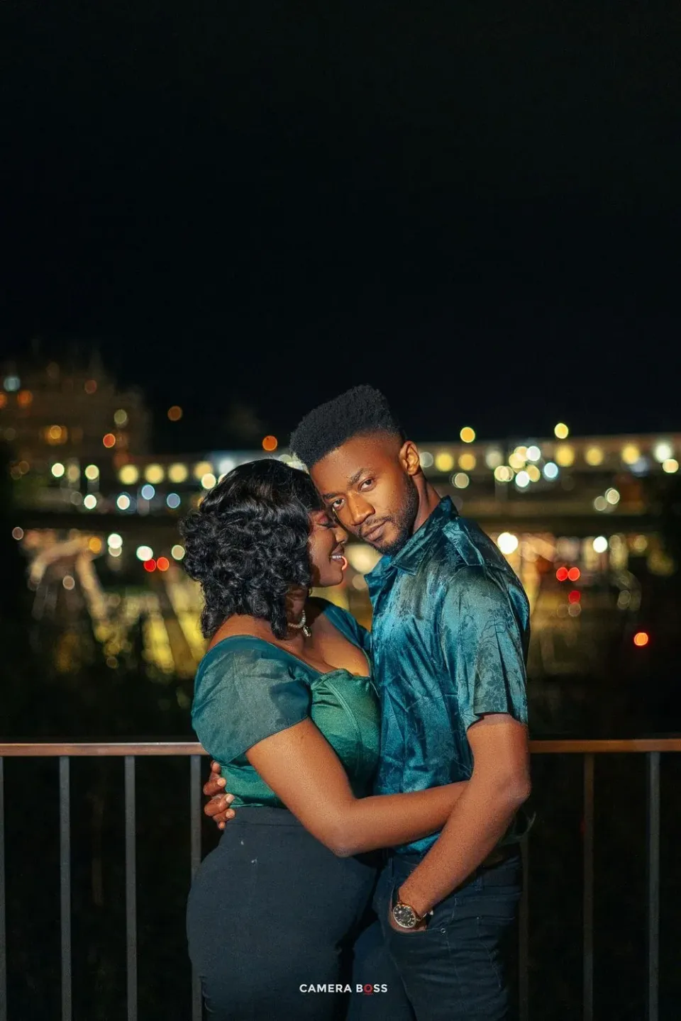 Romantic couple shares intimate moment on balcony at night with city lights twinkling in background.