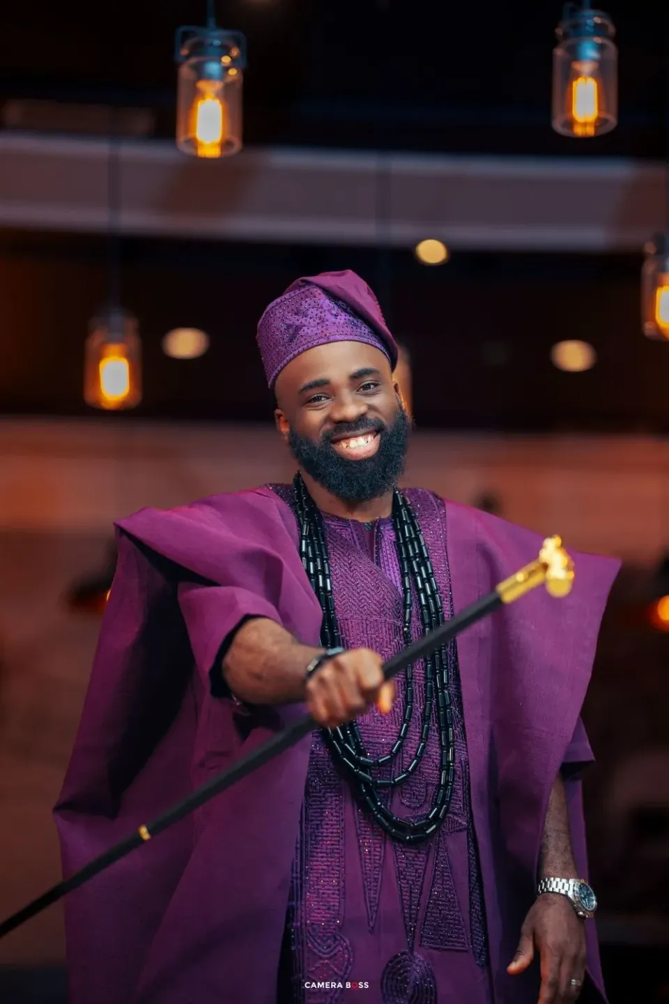 Person wearing vibrant purple traditional Agbada attire with beaded necklaces smiles while holding a staff at indoor event.
