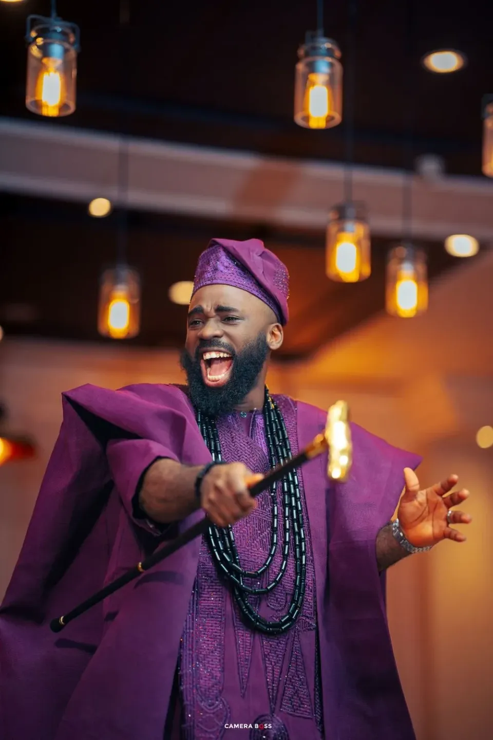 A performer in vibrant purple traditional African attire and beaded necklaces gestures expressively under warm pendant lighting.