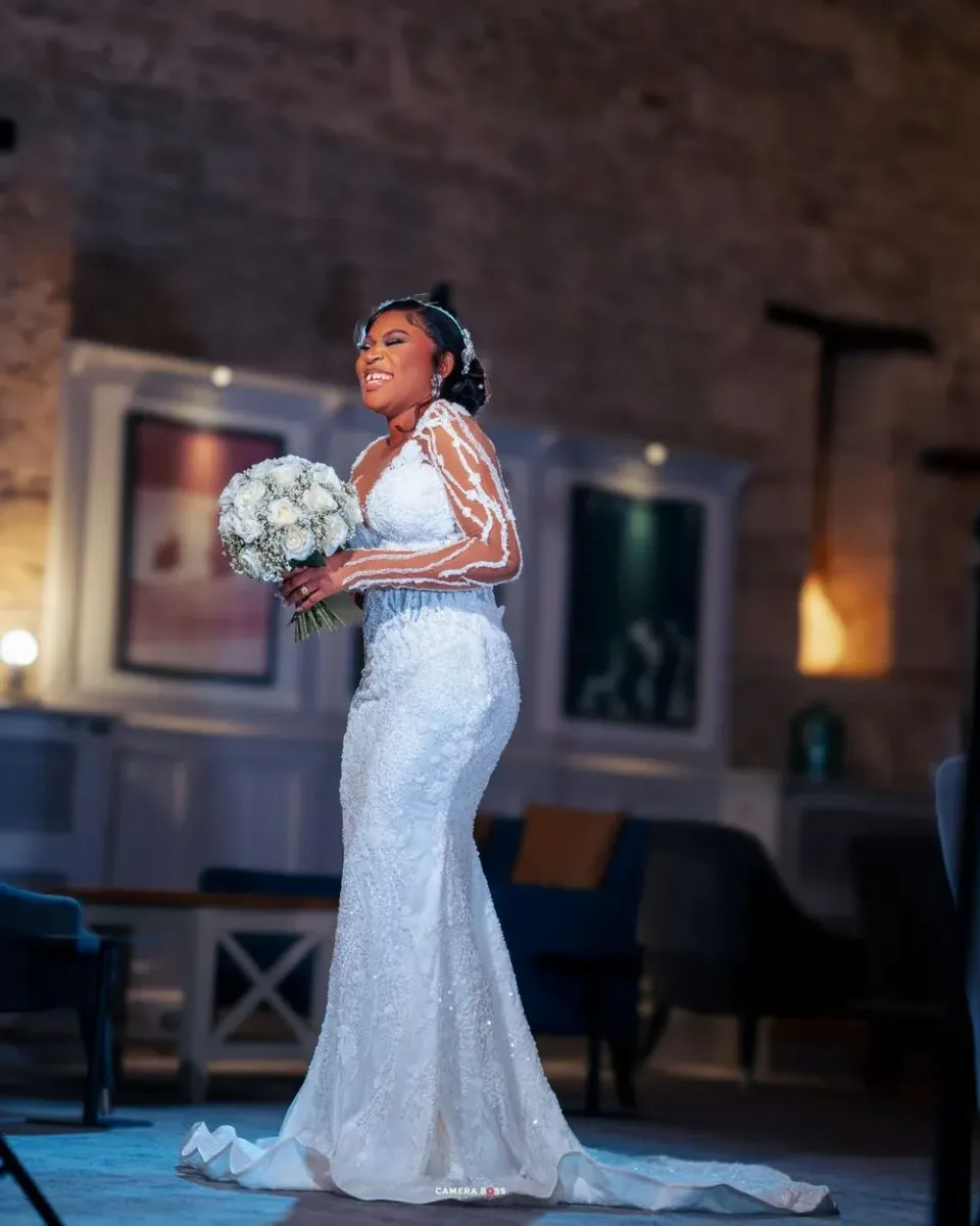 Beautiful bride in lace mermaid wedding gown with long sleeves holds white bouquet while posing in dimly lit venue.