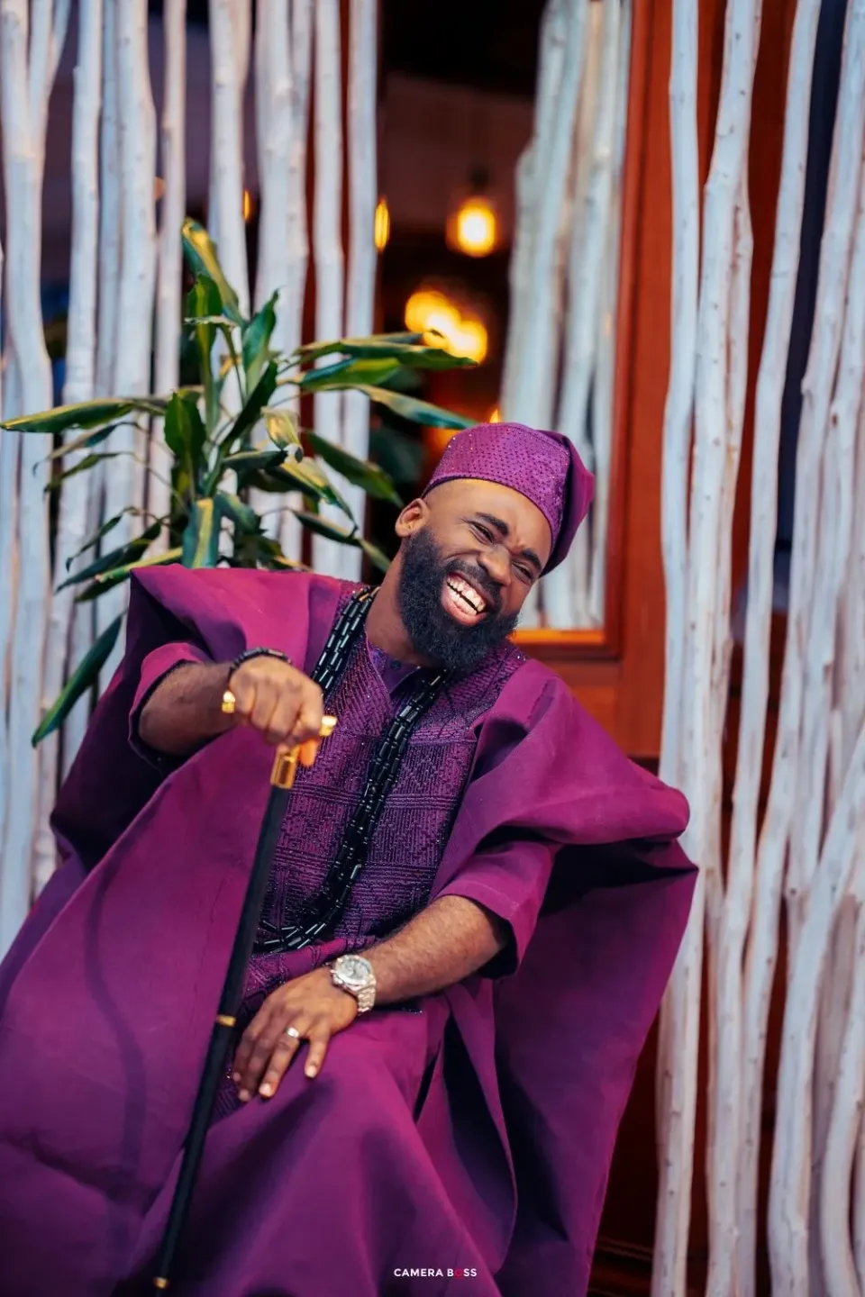 A person in a vibrant purple traditional agbada outfit and matching cap laughs joyfully while standing against a white backdrop.