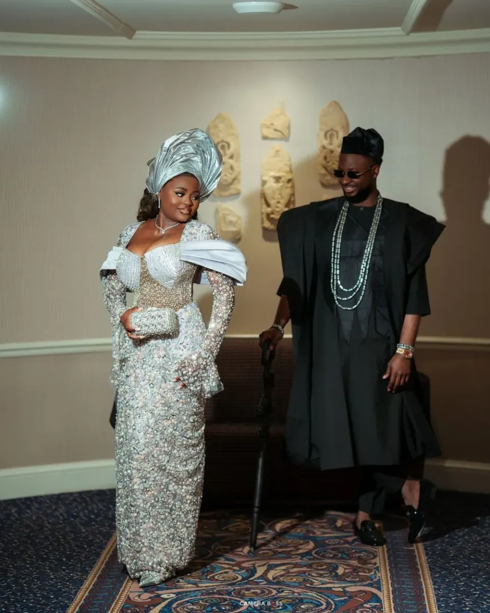 Couple in elegant Nigerian traditional attire, woman in silver gele and lace dress, man in black agbada with beaded necklace.