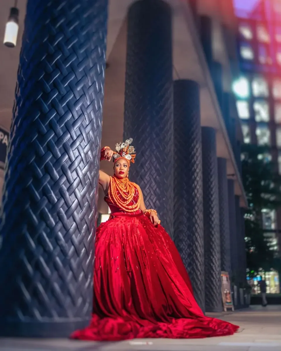 A person in a flowing red gown and ornate headpiece poses dramatically against tall dark textured columns with city lights behind.