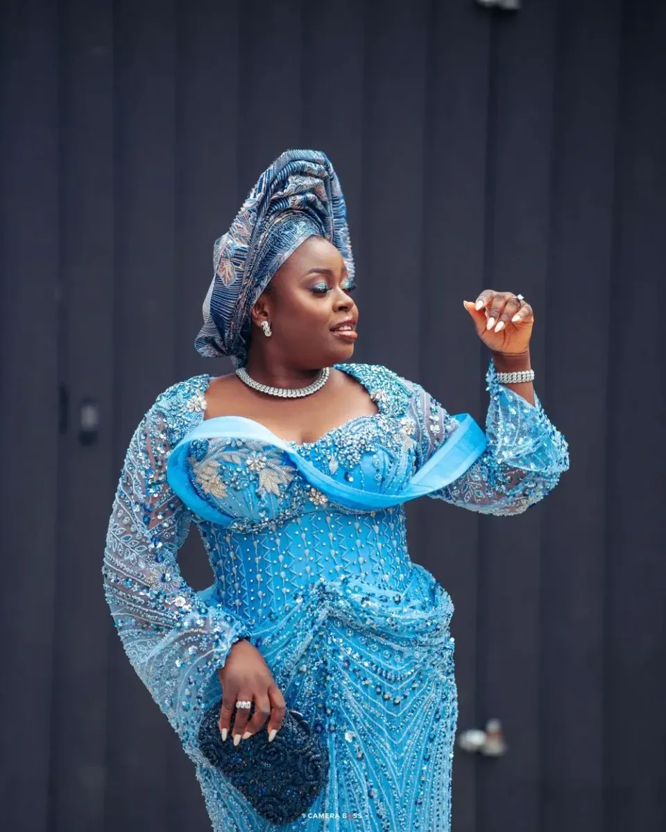 A woman poses gracefully in a sparkling sky blue traditional dress with matching headwrap and pearl necklace.