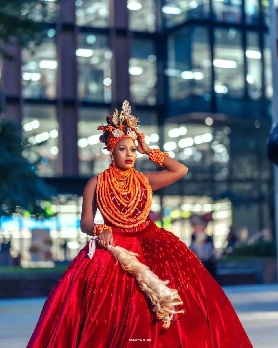 A dancer in a dramatic red ball gown and orange beaded necklaces poses elegantly against a modern glass building backdrop.