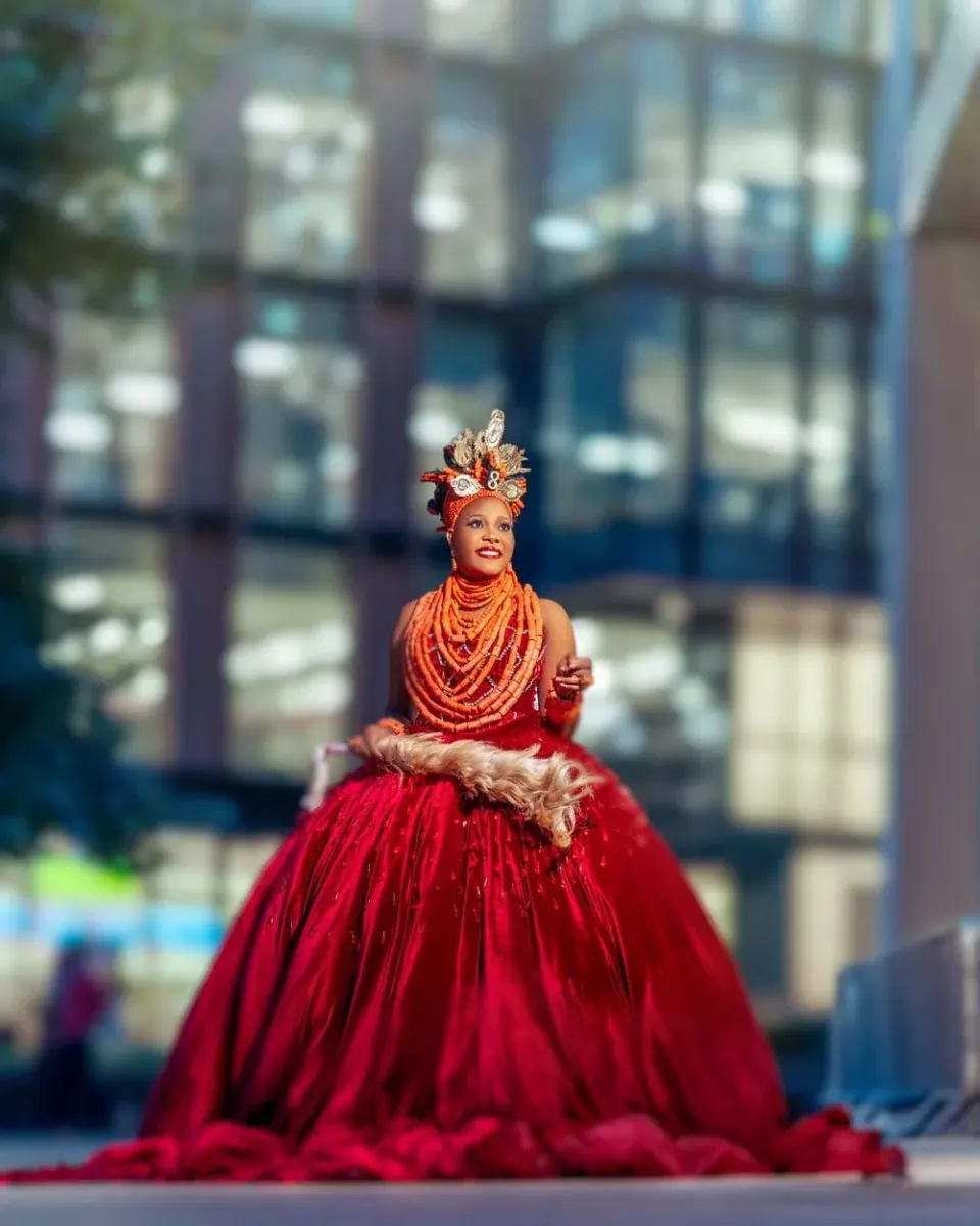 Artistic performer in vibrant orange and red ball gown poses elegantly against modern glass building background.