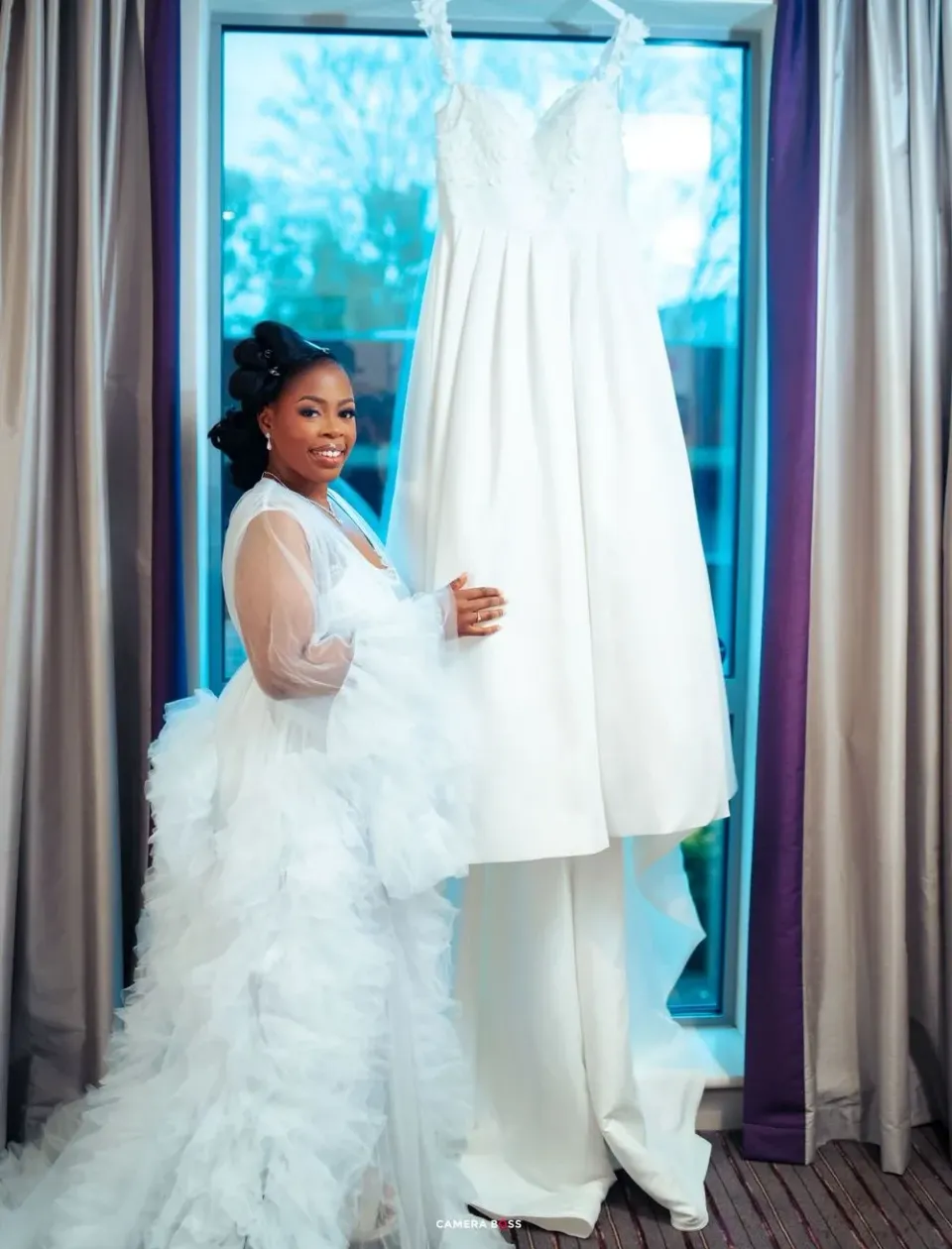 A person in a white robe admires an elegant white wedding dress hanging by a window with teal-colored outdoor scenery.