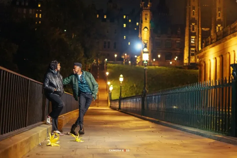 Two people walk together at night along an illuminated path with city buildings and streetlights in the background.